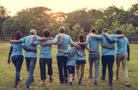 A diverse group of people wearing blue T-shirts walk with their arms around each other away from the observer in the direction of a wood at dusk.